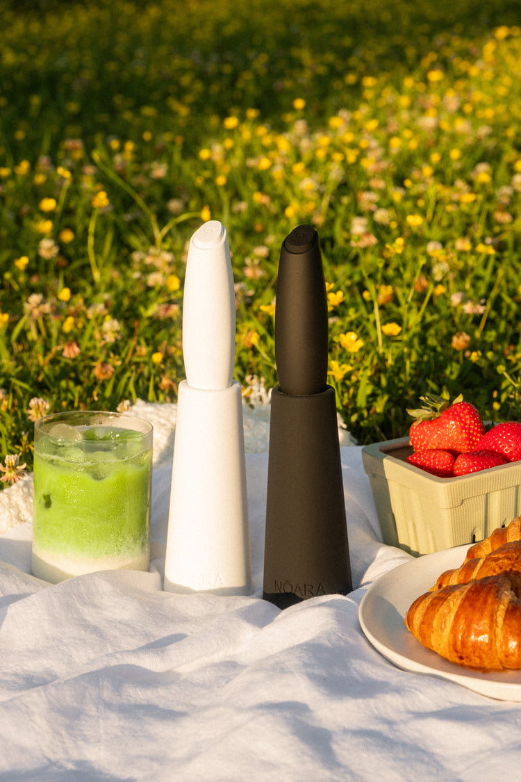 White and black NŌARA frother and silicone stand sets styled outdoors with iced matcha, strawberries, and croissants.